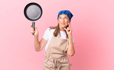 Güzel orta yaşlı bir kadın mutlu bir şekilde gülümsüyor ve hayal kuruyor ya da şüphe ediyor ve elinde bir tava tutuyor. Şef konsepti