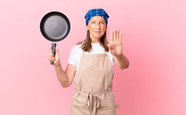 Orta yaşlı bir kadın ciddi görünüyor. Avuç içini gösteriyor. El hareketi yapıyor ve elinde bir tava tutuyor. Şef konsepti