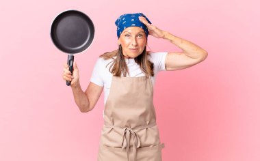 Oldukça orta yaşlı bir kadın stresli, endişeli ya da korkmuş, elleri başının üstünde ve elinde bir tava tutuyor. Şef konsepti