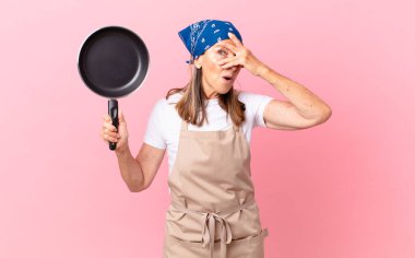 Oldukça orta yaşlı bir kadın şok olmuş, korkmuş ya da korkmuş görünüyor. Yüzü elle kaplanmış ve bir tava tutuyor. Şef konsepti