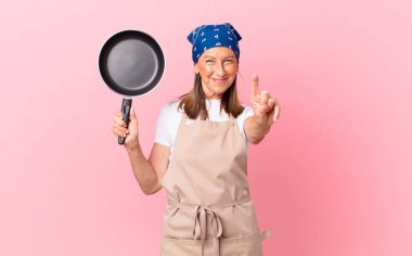 Güzel orta yaşlı bir kadın gülümsüyor ve arkadaş canlısı görünüyor. Bir numarayı gösteriyor ve elinde bir tava tutuyor. Şef konsepti