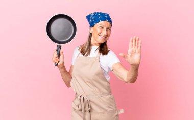 Güzel orta yaşlı bir kadın mutlu bir şekilde gülümsüyor, el sallıyor, sizi selamlıyor ve bir tava tutuyor. Şef konsepti