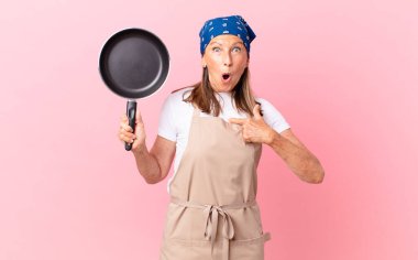 Oldukça orta yaşlı bir kadın ağzı açık, kendini işaret eden ve elinde bir tavayla şaşkın ve şaşırmış görünüyor. Şef konsepti