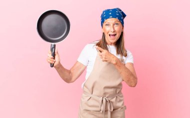 Oldukça orta yaşlı bir kadın heyecanlı ve şaşırmış görünüyor. Bir tarafı işaret ediyor ve elinde bir tava tutuyor. Şef konsepti