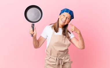 Güzel, orta yaşlı bir kadın, kendine güvenerek gülümsüyor ve elinde bir tava tutuyordu. Şef konsepti