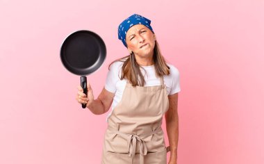 Kafası karışmış, kafası karışmış ve elinde tava tutan orta yaşlı güzel bir kadın. Şef konsepti