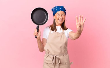 Güzel orta yaşlı bir kadın gülümsüyor ve arkadaş canlısı görünüyor, dört numarayı gösteriyor ve elinde bir tava tutuyor. Şef konsepti