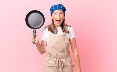 Güzel, orta yaşlı bir kadın mutlu görünüyor ve hoş bir şekilde şaşırmış ve elinde bir tava tutuyor. Şef konsepti