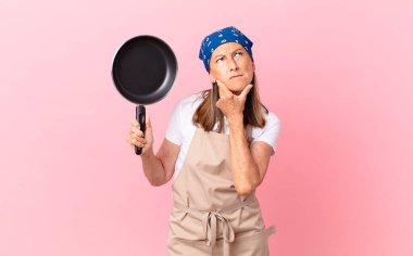 Oldukça orta yaşlı bir kadın düşünüyor, kuşkulu ve kafası karışık hissediyor ve elinde bir tava tutuyor. Şef konsepti