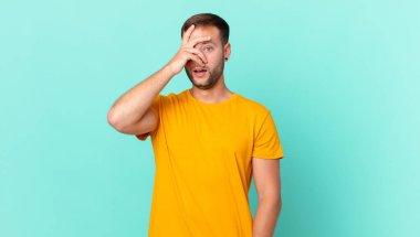 handsome blonde man looking shocked, scared or terrified, covering face with hand