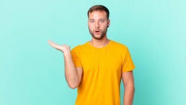 handsome blonde man looking surprised and shocked, with jaw dropped holding an object