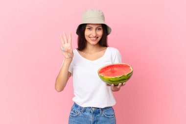 Gülümsüyor ve arkadaş canlısı görünüyor, üç numarayı gösteriyor.