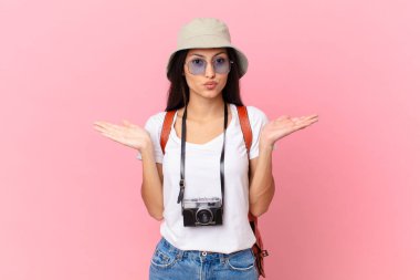 pretty hispanic tourist feeling puzzled and confused and doubting with a photo camera and a hat