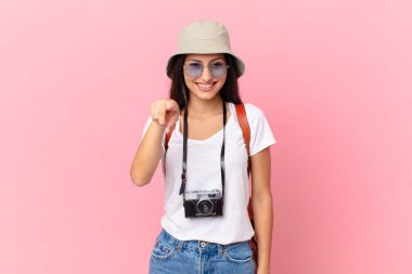 pretty hispanic tourist pointing at camera choosing you with a photo camera and a hat