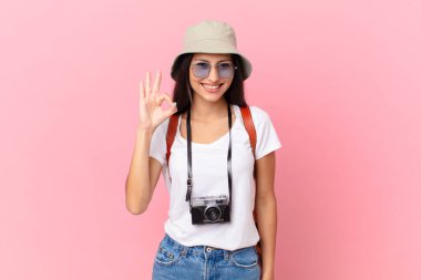 pretty hispanic tourist feeling happy, showing approval with okay gesture with a photo camera and a hat