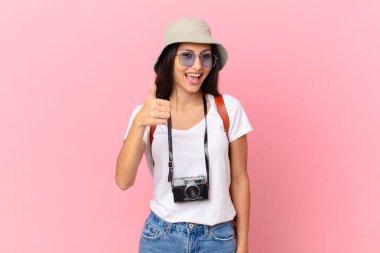 pretty hispanic tourist feeling proud,smiling positively with thumbs up with a photo camera and a hat