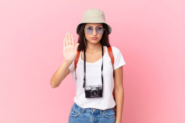 pretty hispanic tourist looking serious showing open palm making stop gesture with a photo camera and a hat