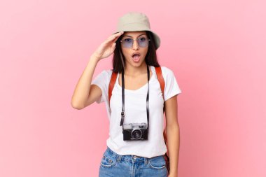 pretty hispanic tourist looking happy, astonished and surprised with a photo camera and a hat