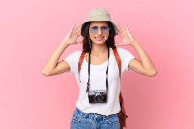 pretty hispanic tourist feeling stressed, anxious or scared, with hands on head with a photo camera and a hat