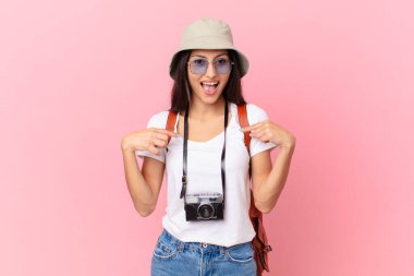 pretty hispanic tourist feeling happy and pointing to self with an excited with a photo camera and a hat