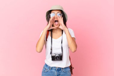 pretty hispanic tourist feeling happy,giving a big shout out with hands next to mouth with a photo camera and a hat