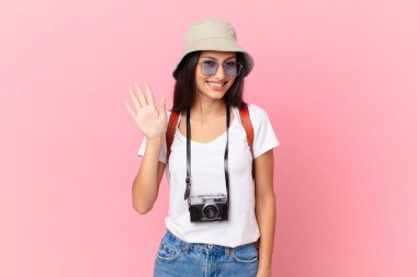 pretty hispanic tourist smiling happily, waving hand, welcoming and greeting you with a photo camera and a hat