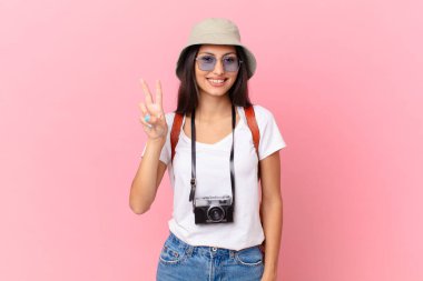 pretty hispanic tourist smiling and looking friendly, showing number two with a photo camera and a hat