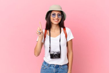 pretty hispanic tourist smiling and looking friendly, showing number one with a photo camera and a hat
