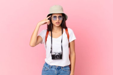 pretty hispanic tourist feeling confused and puzzled, showing you are insane with a photo camera and a hat
