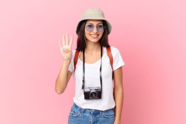 pretty hispanic tourist smiling and looking friendly, showing number four with a photo camera and a hat