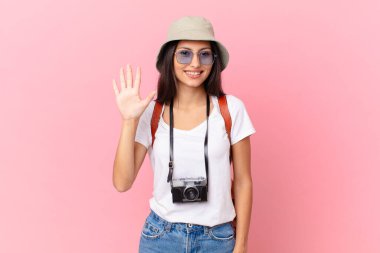 pretty hispanic tourist smiling and looking friendly, showing number five with a photo camera and a hat
