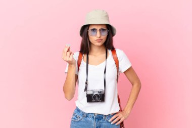 pretty hispanic tourist making capice or money gesture, telling you to pay with a photo camera and a hat