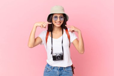 pretty hispanic tourist smiling confidently pointing to own broad smile with a photo camera and a hat