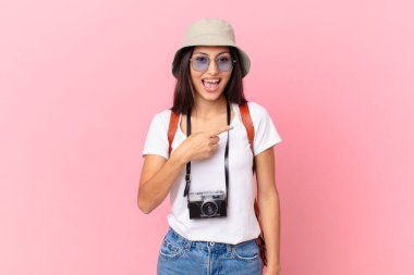 pretty hispanic tourist looking excited and surprised pointing to the side with a photo camera and a hat