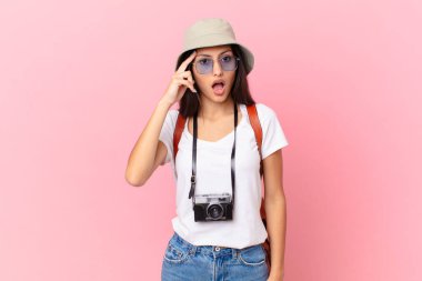 pretty hispanic tourist looking surprised, realizing a new thought, idea or concept with a photo camera and a hat