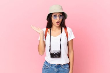 pretty hispanic tourist looking surprised and shocked, with jaw dropped holding an object with a photo camera and a hat