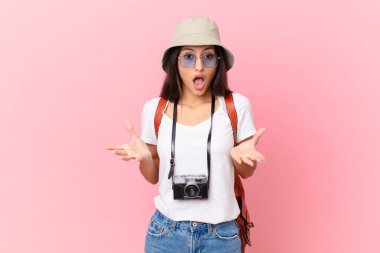 pretty hispanic tourist feeling extremely shocked and surprised with a photo camera and a hat