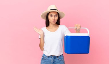 pretty hispanic woman feeling puzzled and confused and doubting and holding a portable refrigerator