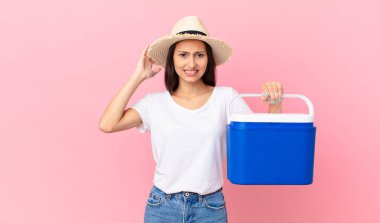 pretty hispanic woman feeling stressed, anxious or scared, with hands on head and holding a portable refrigerator