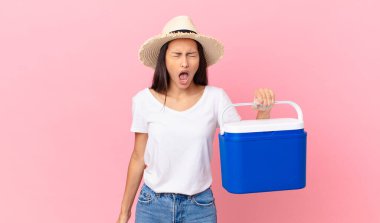 pretty hispanic woman shouting aggressively, looking very angry and holding a portable refrigerator