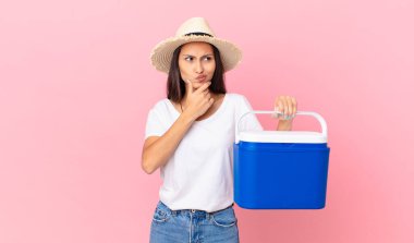 pretty hispanic woman thinking, feeling doubtful and confused and holding a portable refrigerator