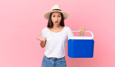 pretty hispanic woman feeling extremely shocked and surprised and holding a portable refrigerator