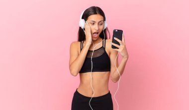 hispanic fitness woman feeling happy, excited and surprised with headphones and a phone