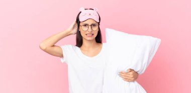 hispanic woman wearing pajamas feeling stressed, anxious or scared, with hands on head and holding a pillow