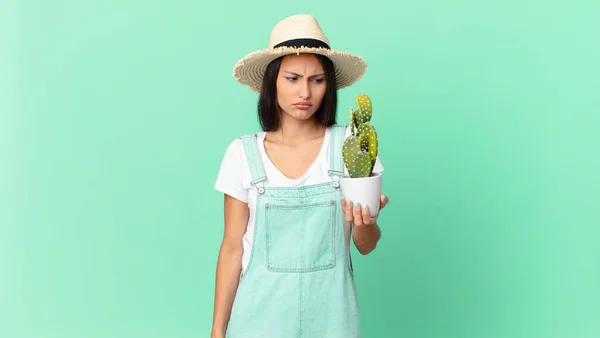 pretty farmer woman feeling sad, upset or angry and looking to the side and holding a cactus