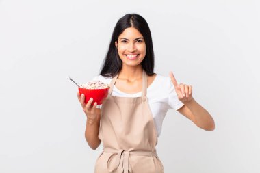 Güzel İspanyol kadın güzel İspanyol şef kadın gülümsüyor ve arkadaş canlısı görünüyor, bir numarayı gösteriyor ve elinde mısır gevreği kasesi var.