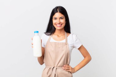 Güzel İspanyol aşçı kadın mutlu bir şekilde gülümsüyor, eli kalçasında, kendinden emin ve elinde süt şişesi var.