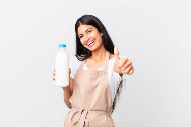 Güzel İspanyol aşçı kadın gururlu, olumlu gülümsüyor. Başparmakları havada ve elinde süt şişesi var.