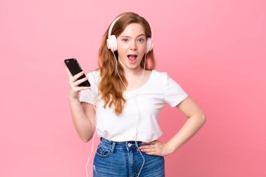 Güzel kızıl saçlı kadın kulaklıklar ve akıllı telefonlarla şok olmuş ya da şaşırmış görünüyor.