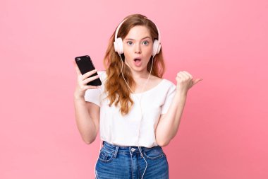 Güzel kızıl saçlı kadın kulaklıklarla ve akıllı telefonlarla şaşkın şaşkın bakıyor.